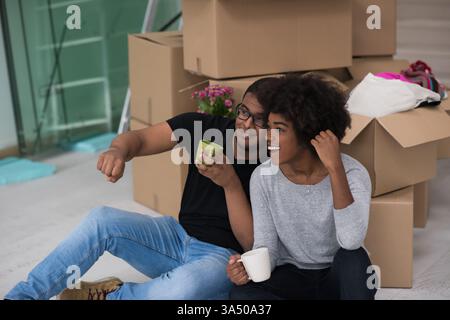 Ein fröhliches, junges afroamerikanisches Paar sitzt auf dem Boden in ihrem neuen Zuhause und genießt Kaffee in Umzugskartons. Diese warme Szene des häuslichen Lebens fängt Umsiedlungen, Wohnsiedlungen und das Glück der Familie ein. Ideal für Immobilien, Umzüge, Wohnkultur und Lifestyle-Marketing. Stockfoto