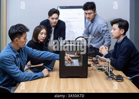 Selbstbewusste chinesische Designer, die einen 3D-Drucker in einem modernen Büro verwenden. Ein kreatives Technologienimage, das sich für Design, Fertigung und Innovation eignet. Hervorragend geeignet für Start-ups im Bereich Technik, Produktentwicklung und Collaboration-Kampagnen. Stockfoto