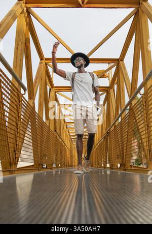 Aus einem niedrigen Winkel steht ein stilvoller Afroamerikaner auf einer Brücke und macht ein Selfie mit seinem Smartphone. Der städtische Rucksack und die Mütze verleihen dem Tourismus und den sozialen Medien ein Gefühl. Perfekt für Lifestyle, Reisen und multiethnische Marketinggrafiken. Stockfoto
