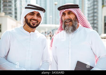 Lächelnde Geschäftsleute aus dem Nahen Osten und Kaukasien, die Kanduras tragen, stehen tagsüber auf einer Straße der Stadt zusammen. Die Szene vermittelt kulturübergreifende Teamarbeit in einem urbanen Umfeld. Geeignet für internationale Geschäfte und visuelle Darstellungen für den Nahen Osten. Stockfoto