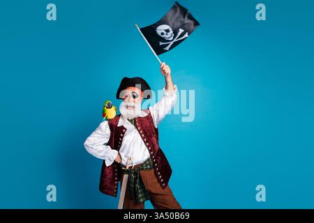 Foto-Porträt des älteren lustigen Piraten Opa Welle Flagge tragen Maskerade Party Kostüm isoliert blau Hintergrund Stockfoto