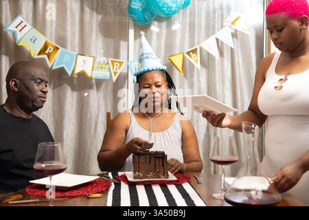 Fröhliche Schwarze Mutter schneidet Kuchen für die Familie, die sie am Esstisch während einer Hausfeier teilen kann. Die Szene strahlt Wärme, Familienleben und Feiern aus. Geeignet für Eltern, Hausleben und Geburtstagsmarketing-Inhalte. Stockfoto