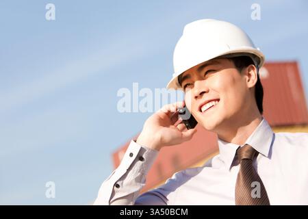 Ein lächelnder chinesischer Geschäftsmann mit einem Schutzhelm plaudert auf seinem Handy an einem belebten Schiffshafen und steht neben Frachtcontainern. Dieses Bild eignet sich ideal für Logistik, Gütertransport und exportorientierte Inhalte, die den modernen Hafenbetrieb hervorheben. Stockfoto