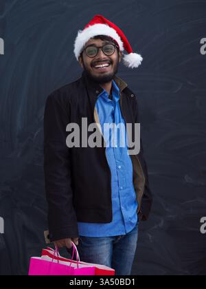 Indischer Mann in festlicher Partykleidung, trägt einen Weihnachtsmann-Hut und hält eine Einkaufstasche vor dunklem Studio-Hintergrund. Das Bild zeigt Weihnachtseinkäufe, Geschenke und Weihnachtsaktionen. Stockfoto