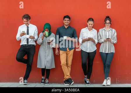 Multiethnische Gruppe von Geschäftsleuten, die während einer Kaffeepause vor einer roten Wand Smartphones nutzen. Die Szene vermittelt Teamwork, Vielfalt und moderne Unternehmenskultur. Stockfoto