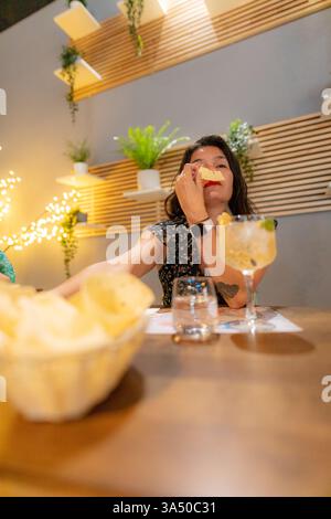 Die hispanische Frau schaut weg, während sie an einem Esstisch in einem Restaurant sitzt. Sie genießt einen Moment der Ruhe in einer modernen, stilvollen Umgebung. Ideal für Gastfreundschaft, Gastronomie oder Lifestyle. Stockfoto