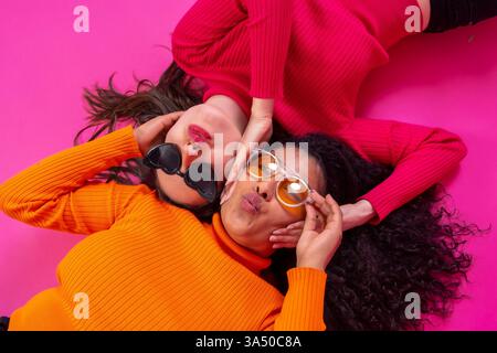 Verschiedene Frauen liegen auf einem rosa Studioboden mit Sonnenbrille und posieren mit Pouty-Lippen. Ein verspieltes, modefokussiertes Bild, das Freundschaft und modernen Stil unterstreicht. Ideal für Lifestyle-, Beauty- und Diversity-Kampagnen. Stockfoto
