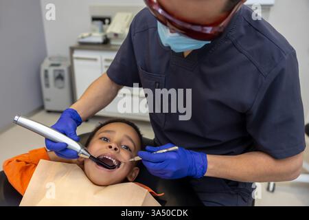 Zahnarzt mit Brille behandelt die Zähne eines Jungen im klinischen Umfeld. Professionelles Gesundheitsbild geeignet für Zahnkliniken, Kinderzahnheilkunde und familiäre Zahnpflege. Klare, informative Visualisierungen für die Gesundheitskommunikation und Patientenaufklärung. Stockfoto