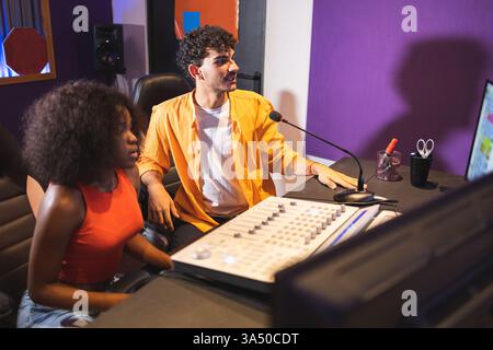 Ein Toningenieur arbeitet mit einer afroamerikanischen Sängerin in einem Kontrollraum des Aufnahmestudios zusammen. Sie arbeiten mit einer computerbasierten Produktionseinrichtung und einer Mischkonsole, um Musik mit professioneller Studioausrüstung zu kreieren. Stockfoto