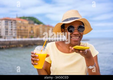 Schwarze ethnische Frau trinkt Orangensaft, während sie in sonniger Umgebung auf ihrem Handy schreibt. Ein Sommer-City-Flair hebt Reisen, Lifestyle und Freizeit hervor. Stockfoto