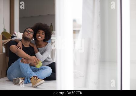 Fröhliches afroamerikanisches Paar entspannt sich auf dem Boden in ihrem neuen Zuhause und genießt Kaffee zwischen Umzugskartons. Der Moment fängt die Besiedlung, den Umzug und das alltägliche Glück ein. Ideal für Lifestyle-, Heim- und Familienmomente. Stockfoto