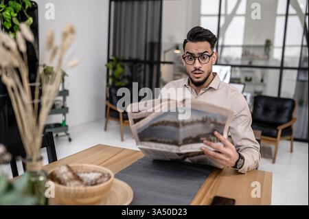 Ein arabischer Mann mit Brille zeigt einen schockierten Ausdruck, während er an einem Tisch im Wohnzimmer liest. Der offene Moment passt zu modernen Lifestyle- und Home Office-Themen. Stockfoto