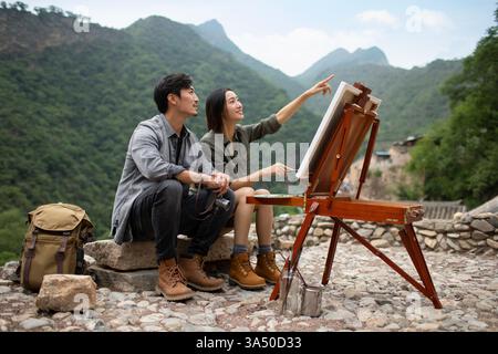 Ein lächelndes chinesisches Paar, das zusammen auf einem Stein auf einem Berggipfel malt. Eine Outdoor-Szene, die Reisen, Natur und Kunst beleuchtet, wobei das Paar im Mittelpunkt steht. Ideal für Lifestyle, Romantik und Kunst im Freien. Stockfoto