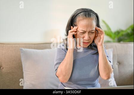 Eine ältere asiatische Frau sitzt zu Hause auf einem Sofa und berührt ihre Schläfe mit einem schmerzhaften Ausdruck. Dieses Gesundheits- und Wellness-Image eignet sich für Artikel und Kampagnen über Migräne, Altenpflege und häusliche Erkrankungen. Stockfoto