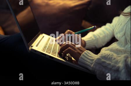 Eine hispanische Frau sitzt zu Hause auf einem Sofa und tippt mit einem Bleistift in der Hand auf einem Laptop. Das gemütliche Wohnzimmer und die warme Beleuchtung sorgen für eine Atmosphäre im Home Office für die Visualisierung von externen Arbeitsumgebungen. Geeignet für digitalen Lifestyle, Produktivität und Arbeit von zu Hause aus. Stockfoto
