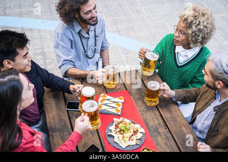 Verschiedene Freunde genießen tagsüber Bier in einer Bar im Freien. Sie lachen, toasten und teilen Tapas in einer lebhaften urbanen Umgebung. Ideal für Kampagnen über Freundschaft, multikulturelle Treffen oder entspanntes Nachtleben in der Stadt. Stockfoto