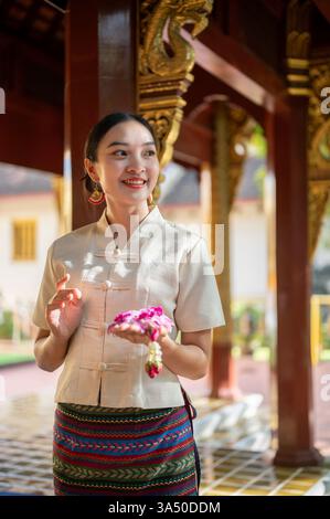 Porträt einer charmanten asiatischen Thai-Frau in traditionellem Thai-Lanna-Kleid mit einer Girlande in der Hand in einem Tempel, die Verdienste macht. Eine authentische Kulturszene aus Thailand, die buddhistische Praktiken und Tempelanlagen hervorhebt. Ideal für Reisen, Kultur und spirituelle Themen mit einer ruhigen, respektvollen Stimmung. Stockfoto