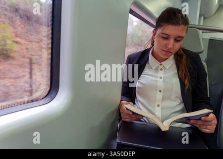 Eine junge Geschäftsfrau liest ein Buch, während sie mit dem Zug unterwegs ist und eine ruhige Reise genießt. Die Szene fängt Fokus, Reisen und das Berufsleben unterwegs ein. Geeignet für Inhalte über Pendeln, Arbeitsreisen und Lernen unterwegs. Stockfoto