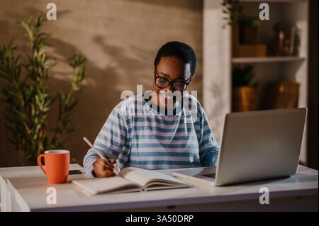 Lächelnde Afroamerikanerin, die von zu Hause aus arbeitet, einen Laptop benutzt und Notizen an einem aufgeräumten Schreibtisch macht. Perfekt für Remote-Arbeiten, Online-Schulungen und Home Office-Themen. Stockfoto