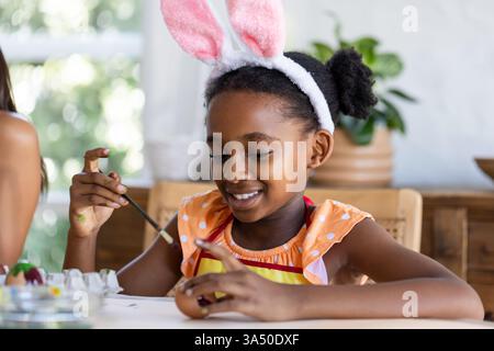 Mit Hasenohren, afroamerikanischem Mädchen, das Osterei malt, glücklich lächelt Stockfoto