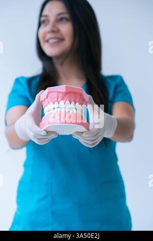 Lächelnde hispanische Zahnärztin in blauen Peelings mit Prothese. Das Porträt steht an einer weißen Wand der Klinik und vermittelt Freundlichkeit und Professionalität. Ideal für Zahnkliniken, Zahnmedizin und Gesundheitsmarketing. Stockfoto