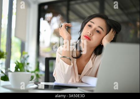 Überarbeitete Millennials asiatische Geschäftsfrau an ihrem Schreibtisch im Büro, die müde und gestresst aussah. Die Szene vermittelt Burnout und Energiemangel an einem modernen Arbeitsplatz. Geeignet für Artikel über Arbeitsstress, Produktivität und Work-Life-Balance. Stockfoto