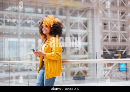 Seitenansicht einer Afroamerikanerin auf einer Stadtstraße, die bei Tageslicht lächelt, während sie auf einem Mobiltelefon surft. Modernes, urbanes Lifestyle-Image, ideal für mobile Technologie, soziale Medien und das vernetzte Leben im Alltag. Stockfoto