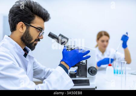 Indischer Wissenschaftler in Labormantel und Handschuhen sitzt mit einem Mikroskop an einem Tisch, während eine Kollegin mit einer Pipette hilft. Das Bild vermittelt die Zusammenarbeit im Labor. Perfekt für die Bereiche Forschung, Biotechnologie und wissenschaftliche Teamarbeit. Stockfoto