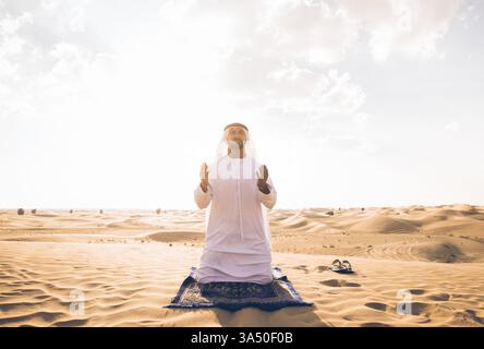 Der arabische Mann in einem traditionellen Kandura kniet tagsüber auf einem Teppich in der Wüste im Gebet. Die Szene vermittelt Hingabe und spirituelle Praxis in einer nahöstlichen Umgebung. Geeignet für Themen zum Islam, Ramadan und religiöse Traditionen in den VAE. Stockfoto