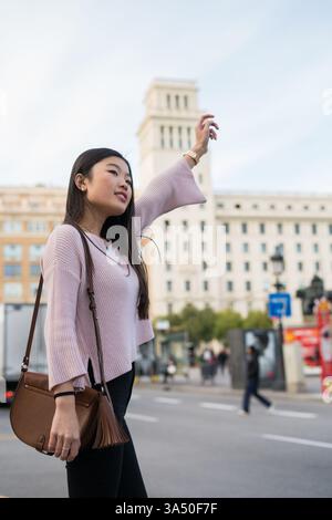 Lächelnde asiatische Frau, die auf einem Bürgersteig der Stadt steht, ruft ein Taxi auf einer belebten Straße. Die urbane Szene reflektiert das Stadtleben und die Verkehrsmittel mit einer kosmopolitischen Atmosphäre. Ideal für Reisen, Lifestyle und Stadtbranding. Stockfoto