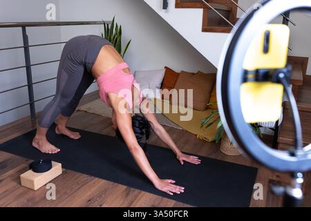 Ein junger, Wellness-orientierter Yogalehrer demonstriert die nach unten gerichtete Hundehaltung während eines Online-Yoga-Kurses, der aus einem Wohnzimmer gestreamt wird. Dieses Bild wurde mit einem Mobiltelefon auf einem Stativ für einen Live-Stream gefilmt und eignet sich ideal für Heimtrainings, virtuelle Yoga-Inhalte und Fitness-Kampagnen in sozialen Medien. Stockfoto