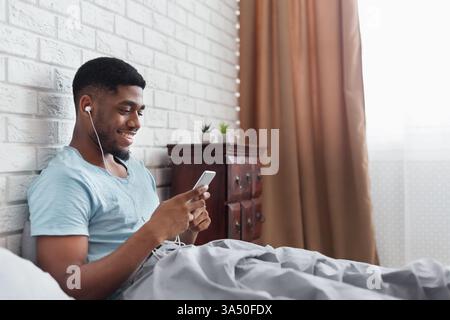 Die entspannte Schlafzimmerszene zeigt einen jungen Schwarzen Mann, der Musik auf einem Smartphone im Bett genießt. Ideal für Lifestyle, Technik und Wellness mit gemütlichem Ambiente und Kopierraum. Stockfoto