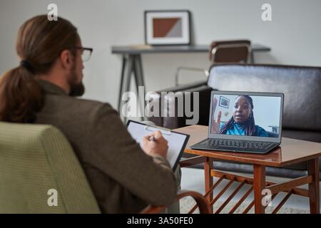 Schwarze Patientin mit Online-Konsultation, die mit einem männlichen kaukasier-Psychologen spricht und Notizen schreibt, während sie in der Klinik auf dem Stuhl sitzt Stockfoto