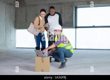 Ein lächelndes Paar aus dem Mittleren Osten und Schwarz steht mit einem Tablet, während ein männlicher kaukasischer Ingenieur sich hockt, um die Pläne für das Haus zu zeigen. Zeigt die Zusammenarbeit im Wohnungsbau und bei der Architekturplanung. Ideal für Immobilien, Wohnbau, Design und familienorientiertes Marketing. Stockfoto