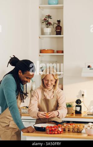 Glückliches, multirassisches Ehepaar, das zu Hause in einer hellen Küche kocht. Sie brechen ein Ei in eine Pfanne, um eine hausgemachte Mahlzeit zuzubereiten und teilen einen heiteren Moment. Eine warme Lifestyle-Szene, in der verschiedene Beziehungen und tägliche Kochkünste hervorgehoben werden. Stockfoto