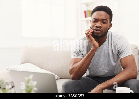 Junger afroamerikanischer Geschäftsmann, der an Zahnschmerzen leidet, während er am Laptop arbeitet, zu Hause auf der Couch sitzt, im Kopierraum ist Stockfoto