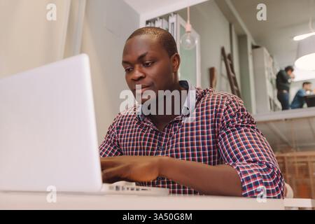 Seriöser Schwarzer in einem karierten Hemd, der an einem Laptop an einem Tisch im Büro arbeitet. Ideal für Unternehmen, Technologie und Coworking. Stockfoto