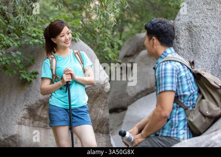 Lächelnde Chinesin mit einem Wanderstock plaudert mit ihrem Mann, während sie während einer Wanderung auf einem Felsen im Wald sitzt. Die Szene fängt einen fröhlichen Moment in der Natur und Outdoor-Erholung in China ein. Geeignet für Reisen, Wandern, Paare und Ökotourismus. Stockfoto