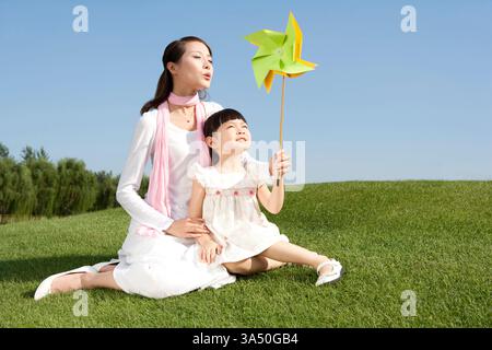 Chinesische Mutter und Tochter spielen zusammen in einem Park und bewundern eine Spielzeugwindmühle. Dieses fröhliche Lifestyle-Bild zeigt Familienbeziehungen, Elternschaft und Freizeitaktivitäten im Freien. Stockfoto