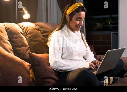 Lächelnde hispanische Frau, die sich zu Hause entspannt, mit gelbem Stirnband und weißem Strickpullover, während sie von einem gemütlichen Wohnzimmer aus an einem Laptop arbeitet. Ein warmes, modernes Home-Office-Ambiente, geeignet für Fernarbeit, digitale Nomaden oder Lifestyle-Inhalte. Hervorragend geeignet für Funktionen zur Vereinbarkeit von Beruf und Privatleben und zur Produktivität zu Hause. Stockfoto