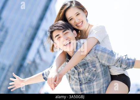 Glückliches junges chinesisches Paar, das einen unbeschwerten Moment im Freien in sonniger urbaner Umgebung genießt. Die Szene fängt Jugend, Romantik und Zweisamkeit mit lässiger Kleidung und einer verspielten Atmosphäre ein. Ideal für Dating, Beziehungen und Kampagnen zum modernen Stadtleben. Stockfoto