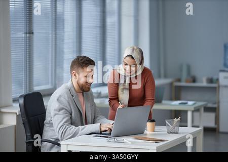 Gemischte muslimische Frau mit Kopftuch, die neben einem männlichen kaukasischen Kollegen steht und über Finanzdaten über Laptops spricht, die im Büro zusammenarbeiten Stockfoto