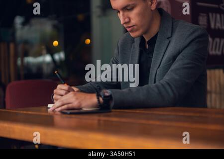 Ein professioneller Mann in formeller Kleidung sitzt an einem hölzernen Kaffeetisch und macht Notizen in einem Notizbuch mit einem Marker. Die Szene vermittelt Geschäftsfokus und Planung in einem zwanglosen Arbeitsumfeld. Hervorragend geeignet für Unternehmensinhalte, Startup-Inhalte oder unternehmerische Initiative. Stockfoto