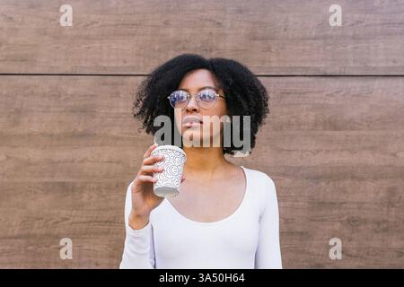 Porträt einer spanischen Frau, die eine Kaffeetasse zum Mitnehmen hält und gegen eine Holzwand blickt. Ideal für den urbanen Lebensstil, Kaffee- oder Café-Konzepte und morgendliche Routinen. Natürliches Tageslicht sorgt für Wärme und eine besinnliche Stimmung. Stockfoto