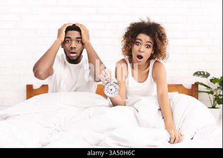 Emotionales afroamerikanisches Paar sitzt auf dem Bett, überrascht vom Wecker, der einen späten und geschäftigen Morgen signalisiert. Dieses Bild eignet sich gut für Lifestyle, Humor und morgendliche Routinethemen. Stockfoto