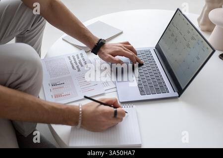 Zugeschnittene Aufnahme eines Mannes aus dem Nahen Osten, der an Berichten an einem Tisch im Wohnzimmer arbeitet. Das Home Office legt Wert auf Konzentration und analytische Arbeit. Perfekt für Remote-Arbeiten, Analysen und Geschäftsplanungsthemen. Stockfoto