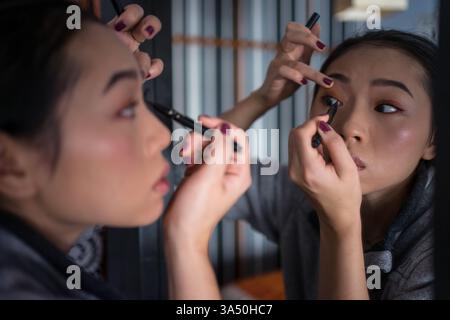 Asiatische Frau, die vor einem Spiegel Eyeliner aufträgt und sich mit Make-up fertig macht. Diese Beauty Shot eignet sich für Make-up-Tutorials, Hautpflege und tägliche Präsentationen zur Vorbereitung. Stockfoto