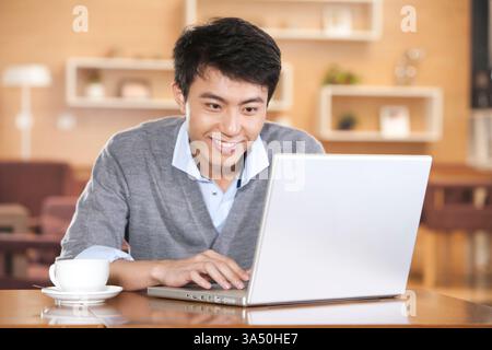 Ein junger Chinese arbeitet an einem Laptop in einem Café, konzentriert sich auf seinen Bildschirm. Die Café-Umgebung und die drahtlose Konnektivität vermitteln Remote-Arbeit und digitale Nomaden-Vibes. Ideal für Inhalte über Online-Produktivität, Notebooks und Coffee-Shop-Kultur. Stockfoto