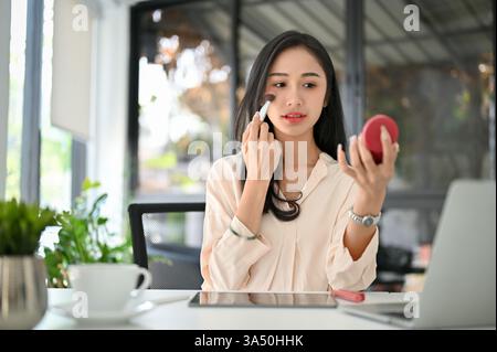 Attraktive Millennials asiatische Geschäftsfrau, die Rouge an ihrem Schreibtisch anwendet. Dieser schöne und modische Moment fängt professionellen Stil und persönliche Pflege am Arbeitsplatz ein. Ideal für Kampagnen zu Schönheit am Arbeitsplatz, Mode und weiblichem Unternehmertum. Stockfoto