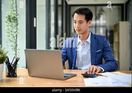 Hübscher asiatischer Geschäftsmann in einem formellen Anzug, der sich in einem modernen Büro auf seinen Laptop konzentriert. Dieses professionelle Porträt eignet sich für Unternehmenskampagnen, Finanz- und Führungskampagnen. Ideal für Geschäfts-, Technologie- und Arbeitsplatzthemen. Stockfoto
